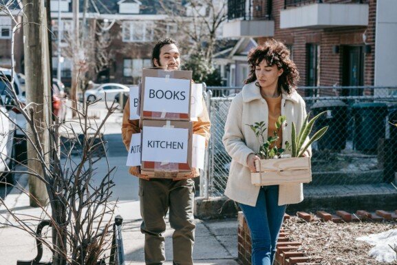 Two people carrying boxes of books on a sidewalk, preparing for their move to the USA.