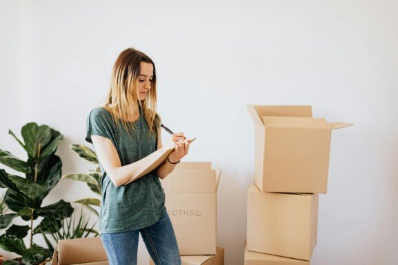 a woman writing surrounded by boxes and taking notes on what to bring in moving abroad