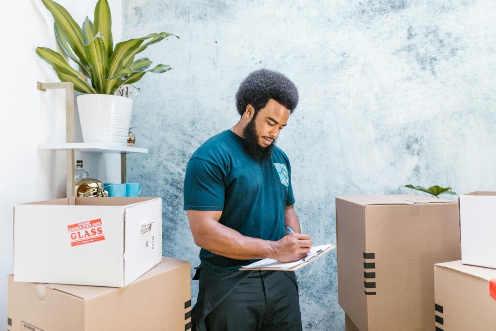 A man stands by boxes, writing on a clipboard, preparing for his move to the US.