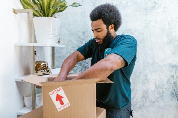 . A man with an afro is packing a box, preparing for his move to the US.